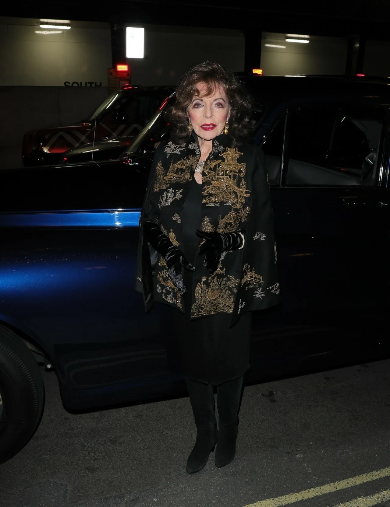 Elegancka kobieta w czarnym płaszczu, ozdobionym wzorzystą szarfą, stoi przed ciemnoniebieskim samochodem nocą na tle miejskiej ulicy.