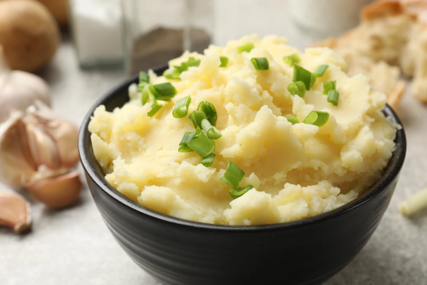 Jak zrobić dobre purée ziemniaczane? Sekretem jest jeden składnik Porcja kremowego puree ziemniaczanego podana w czarnej misce, posypana świeżo posiekanym szczypiorkiem, w tle widoczne ząbki czosnku oraz składniki kulinarne.