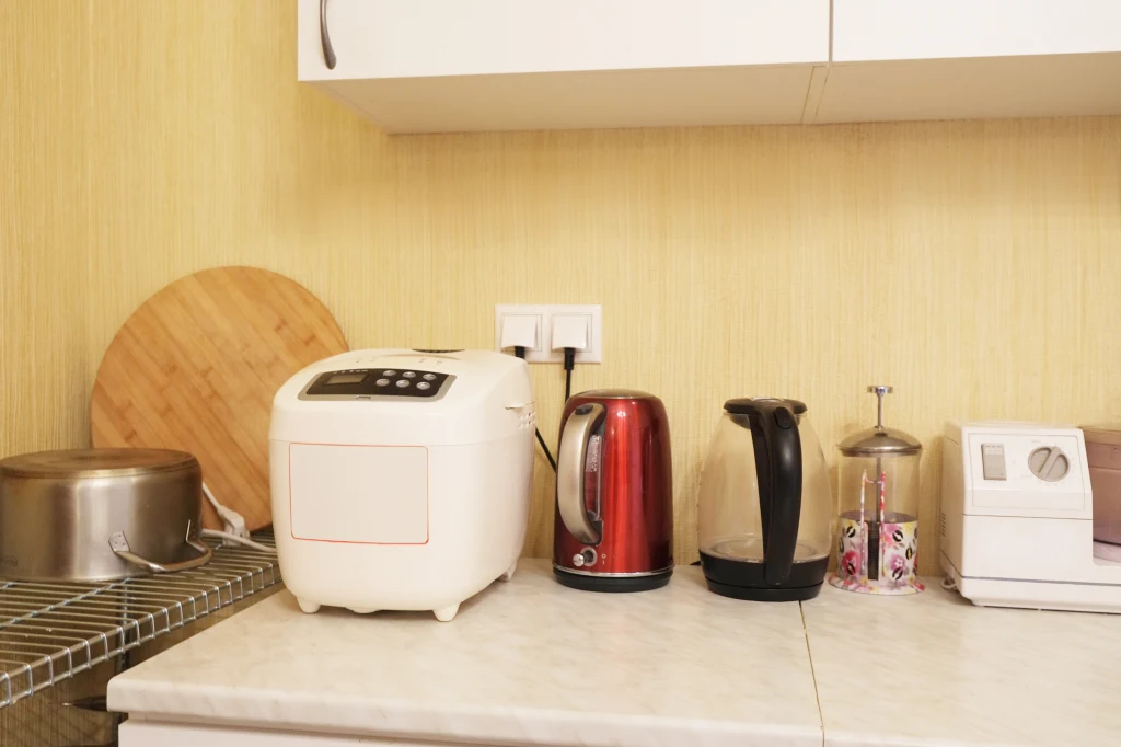 Niektóre urządzenia powinno się wypinać z gniazdka, bo stwarzają zagrożenie pożaru Na blacie kuchennym ustawione są różne sprzęty AGD, takie jak automat do pieczenia chleba, czajnik elektryczny, ekspres do kawy z kapsułkami, a także patelnia i okrągła drewniana deska oparta o ścianę.