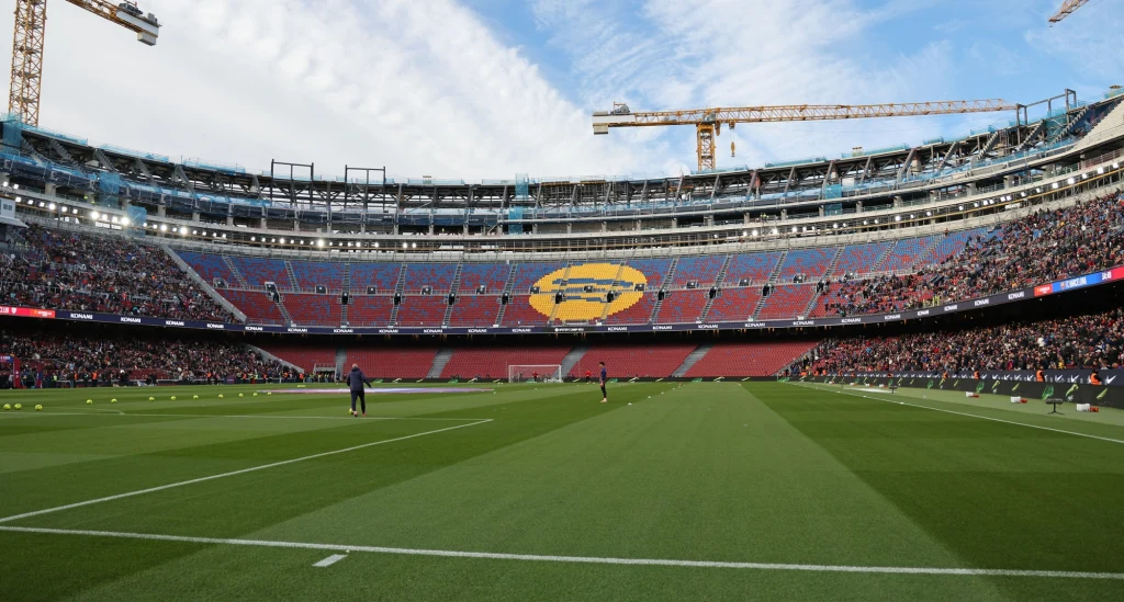Camp Nou Wnętrze dużego stadionu piłkarskiego z zieloną murawą, trybunami częściowo wypełnionymi kibicami oraz charakterystycznym herbem klubu sportowego na centralnych sektorach. Na płycie boiska widoczne są pojedyncze postacie, a nad stadionem unoszą się żura...