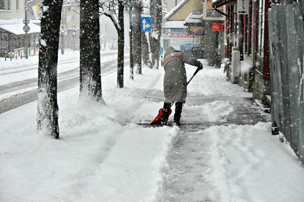 Osoba odśnieżająca chodnik czerwoną łopatą podczas intensywnych opadów śniegu, śnieg pokrywa ulicę, drzewa i budynki po bokach, zimowa sceneria miejska z ograniczoną widocznością.