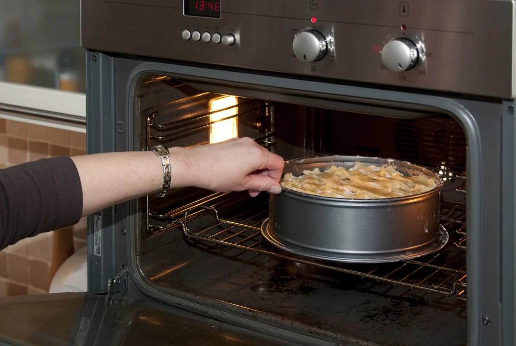 Ręka wkłada tortownicę z ciastem do nagrzanego piekarnika. Wnętrze kuchni oraz panel sterowania piekarnika są widoczne w tle.