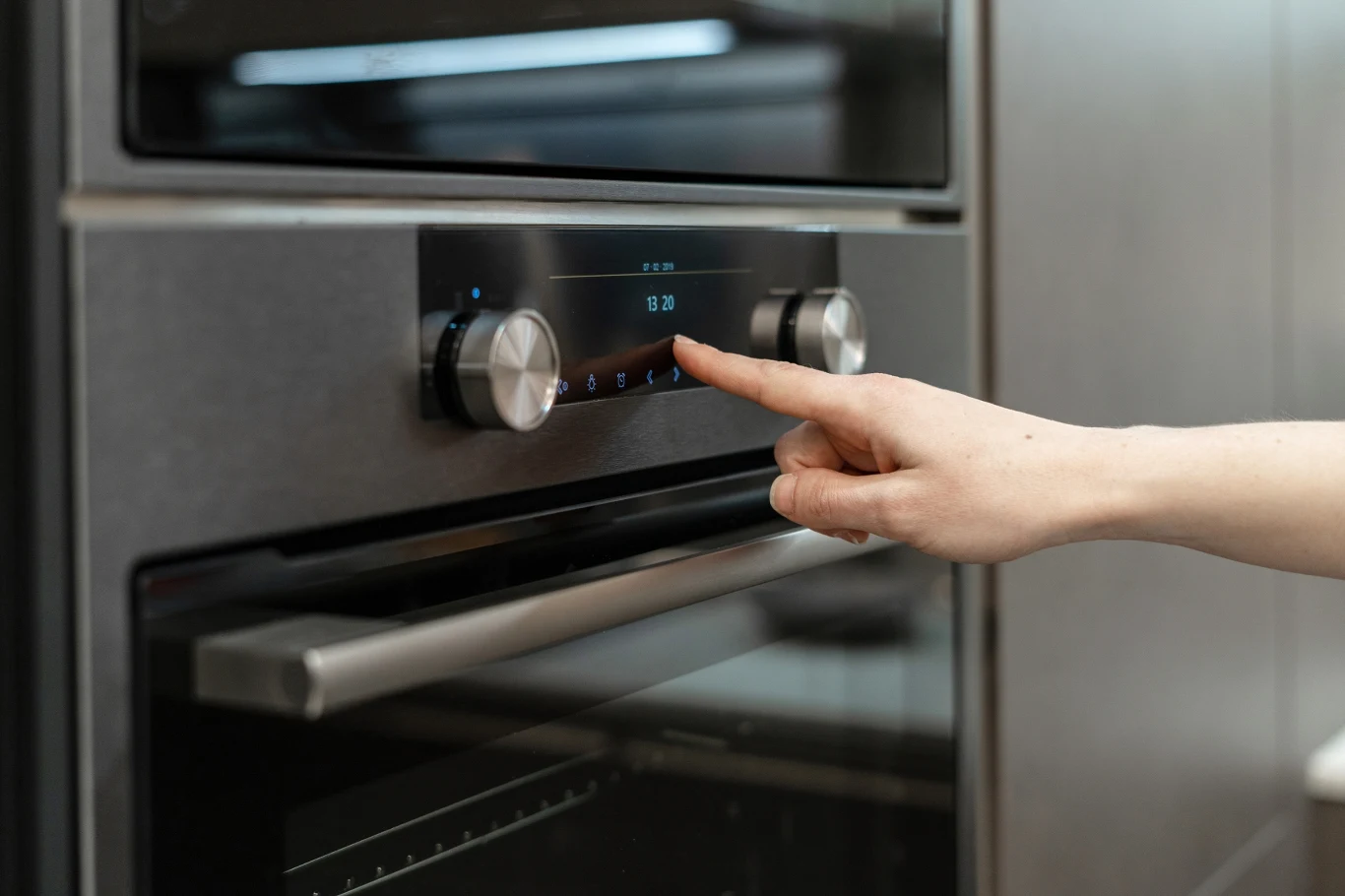 Dłoń dotykająca panel sterujący nowoczesnego piekarnika z elektronicznym wyświetlaczem i pokrętłami, w otoczeniu kuchennej zabudowy.