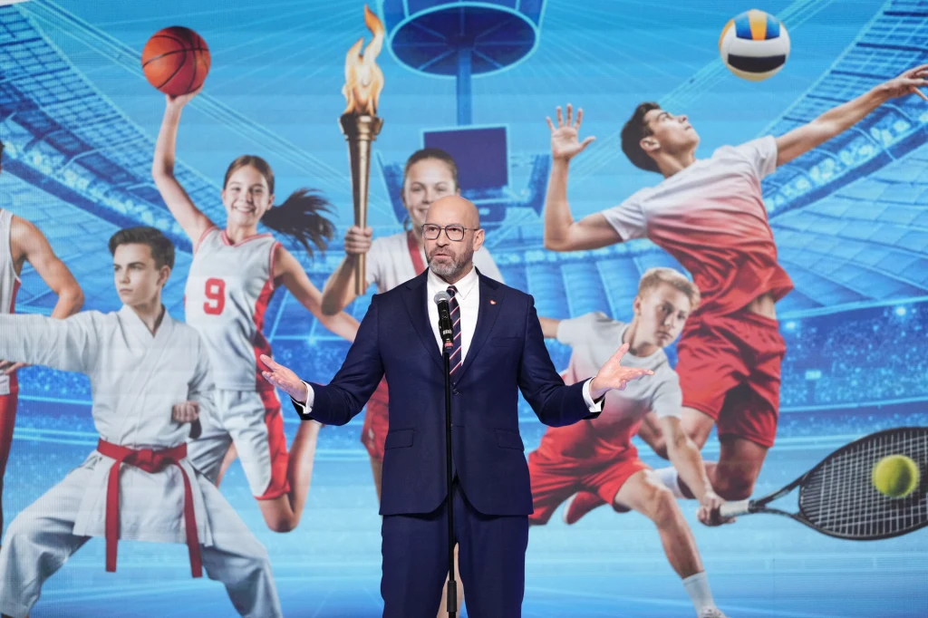 Mężczyzna w garniturze przemawia na tle wielkoformatowej grafiki z młodymi sportowcami uprawiającymi różne dyscypliny, takich jak koszykówka, siatkówka, tenis, karate i lekkoatletyka.