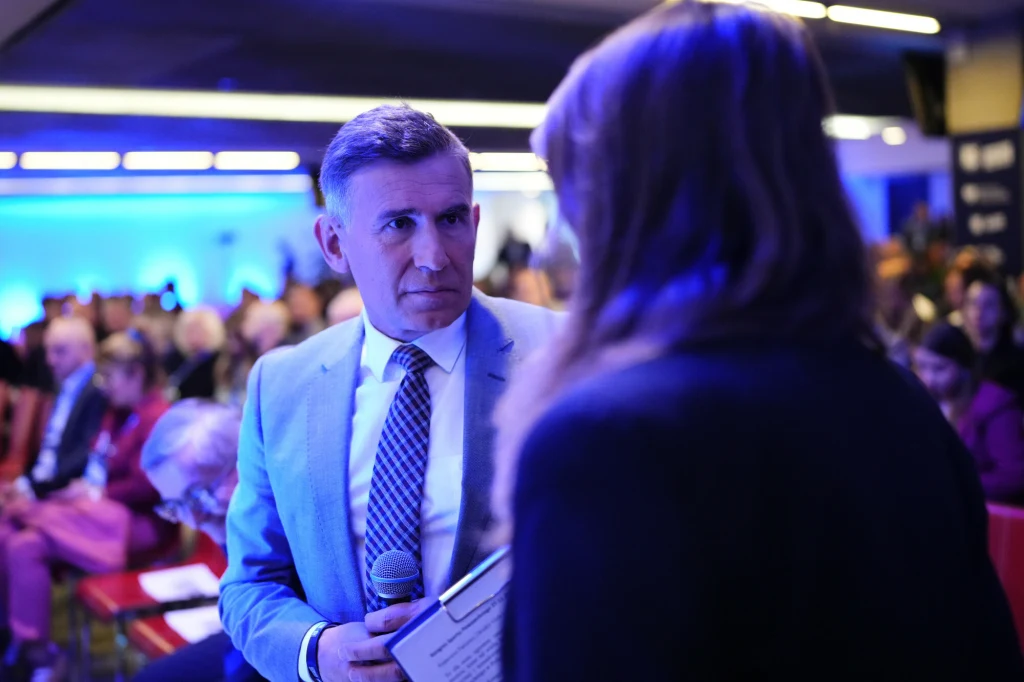 Mężczyzna w garniturze i krawacie trzymający mikrofon oraz teczkę, rozmawiający z kobietą we wnętrzu oświetlonym niebieskim światłem. W tle widoczni ludzie siedzący w rzędach na sali konferencyjnej.