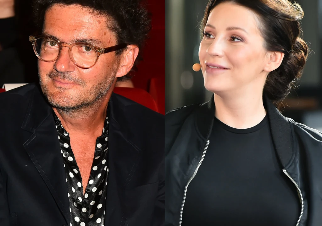 Dwie osoby, mężczyzna z kręconymi włosami w okularach i koszuli w grochy, siedzi w czerwonym fotelu, obok kobiety z upiętymi ciemnymi włosami i lekkim uśmiechem, ubranej na czarno, ze słuchawką przypiętą do policzka.