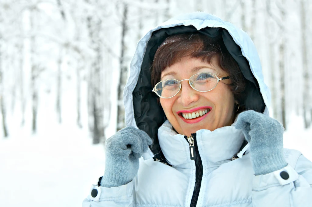 Uśmiechnięta kobieta w okularach i białej, zimowej kurtce z kapturem stoi na tle zaśnieżonego lasu. Trzyma ręce przy kapturze, ubrana jest w szare rękawiczki, wokół widoczne są ośnieżone drzewa.
