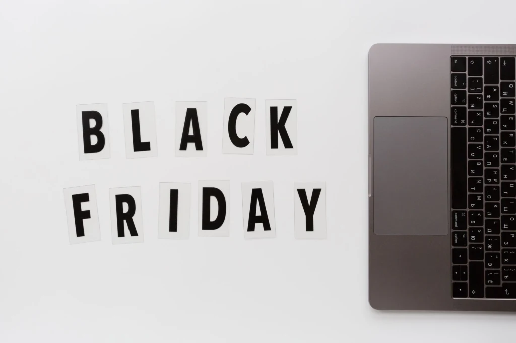 Black Weeks w Media Expert Napis „BLACK FRIDAY” ułożony z czarnych liter na białych kartkach obok fragmentu otwartego laptopa na białym tle.