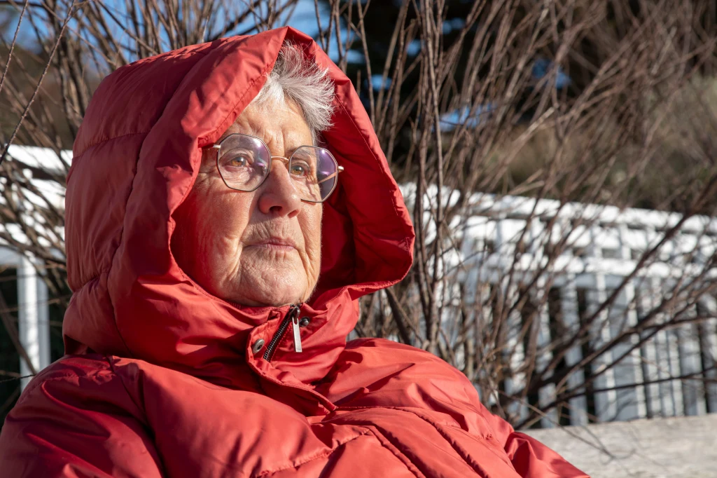 Starsza kobieta w okularach i czerwonej pikowanej kurtce z kapturem, siedząca na ławce na tle bezlistnych gałęzi oraz białego, metalowego ogrodzenia, zamyślona patrząca w dal.