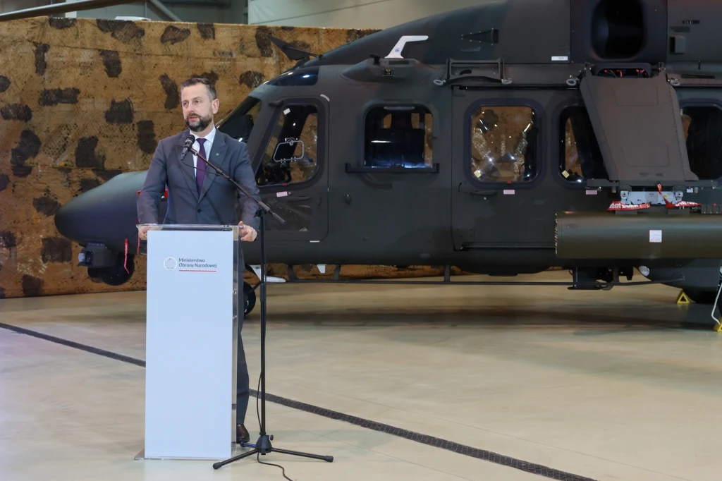 Władysław Kosiniak-Kamysz: AW149 to bardzo dobry śmigłowiec wielozadaniowy, Mężczyzna w garniturze stoi przy mównicy z mikrofonem, za nim znajduje się nowoczesny wojskowy śmigłowiec na tle maskującej siatki.