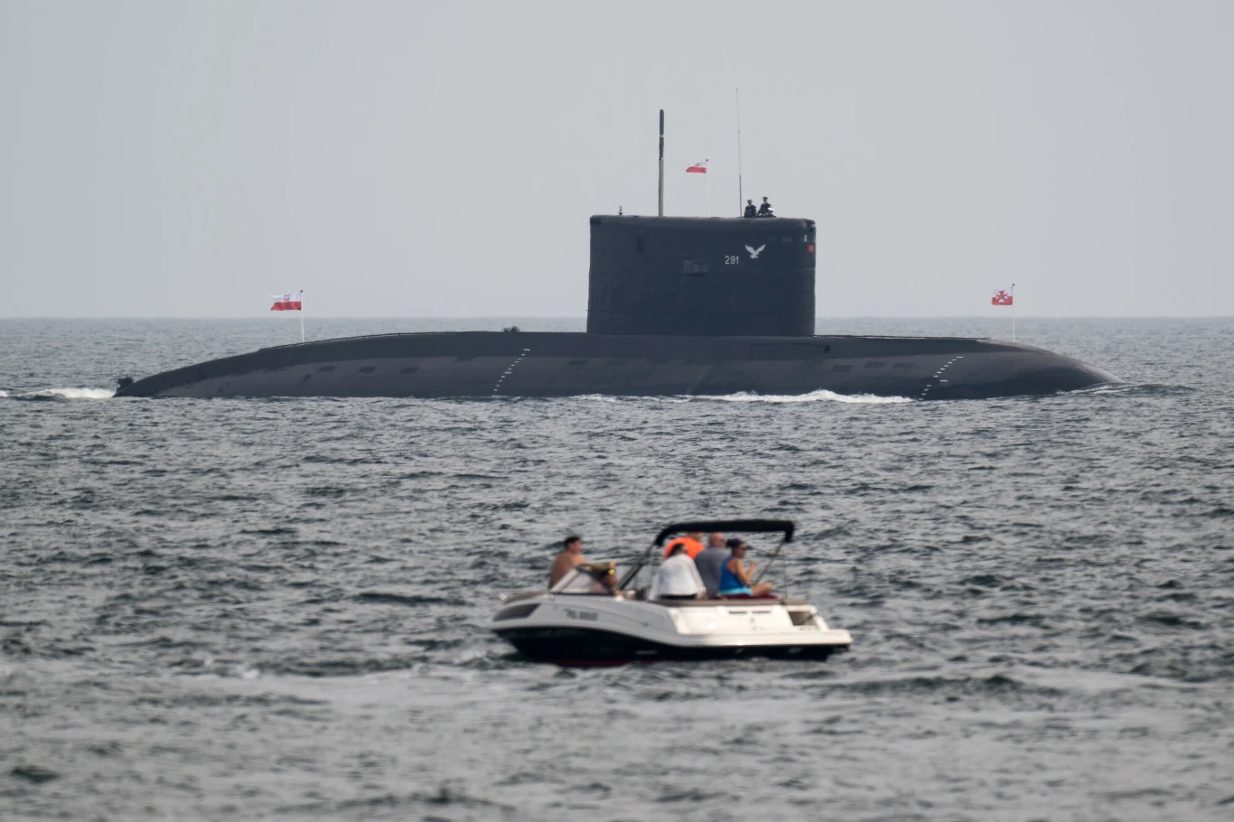 Polska kupi nowe okręty od Szwecji. Media: To zamiana łady na nowe volvo Na wodzie unosi się duży okręt podwodny, częściowo wynurzony, w tle z zamontowanymi flagami, a na pierwszym planie kilku ludzi płynie motorówką.