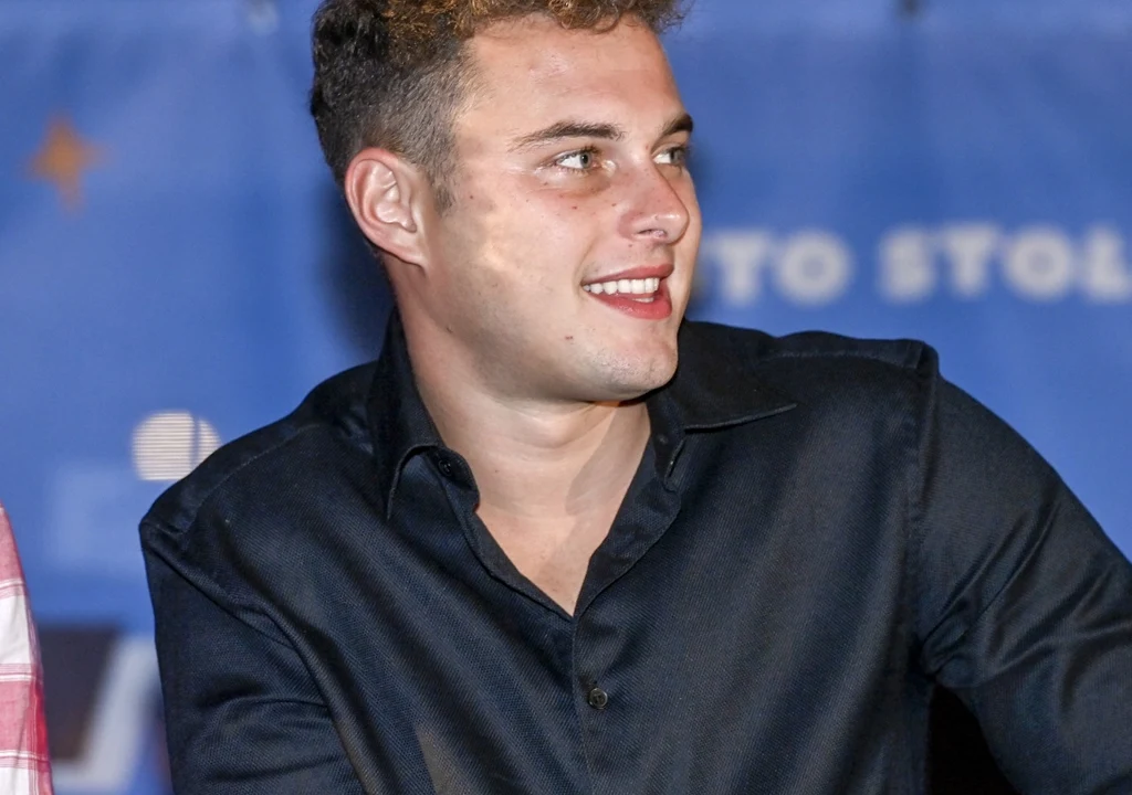 Młody mężczyzna z jasnymi włosami i w ciemnej koszuli siedzi, kierując wzrok w prawo, uśmiechnięty, na niebieskim tle z jasnymi napisami.