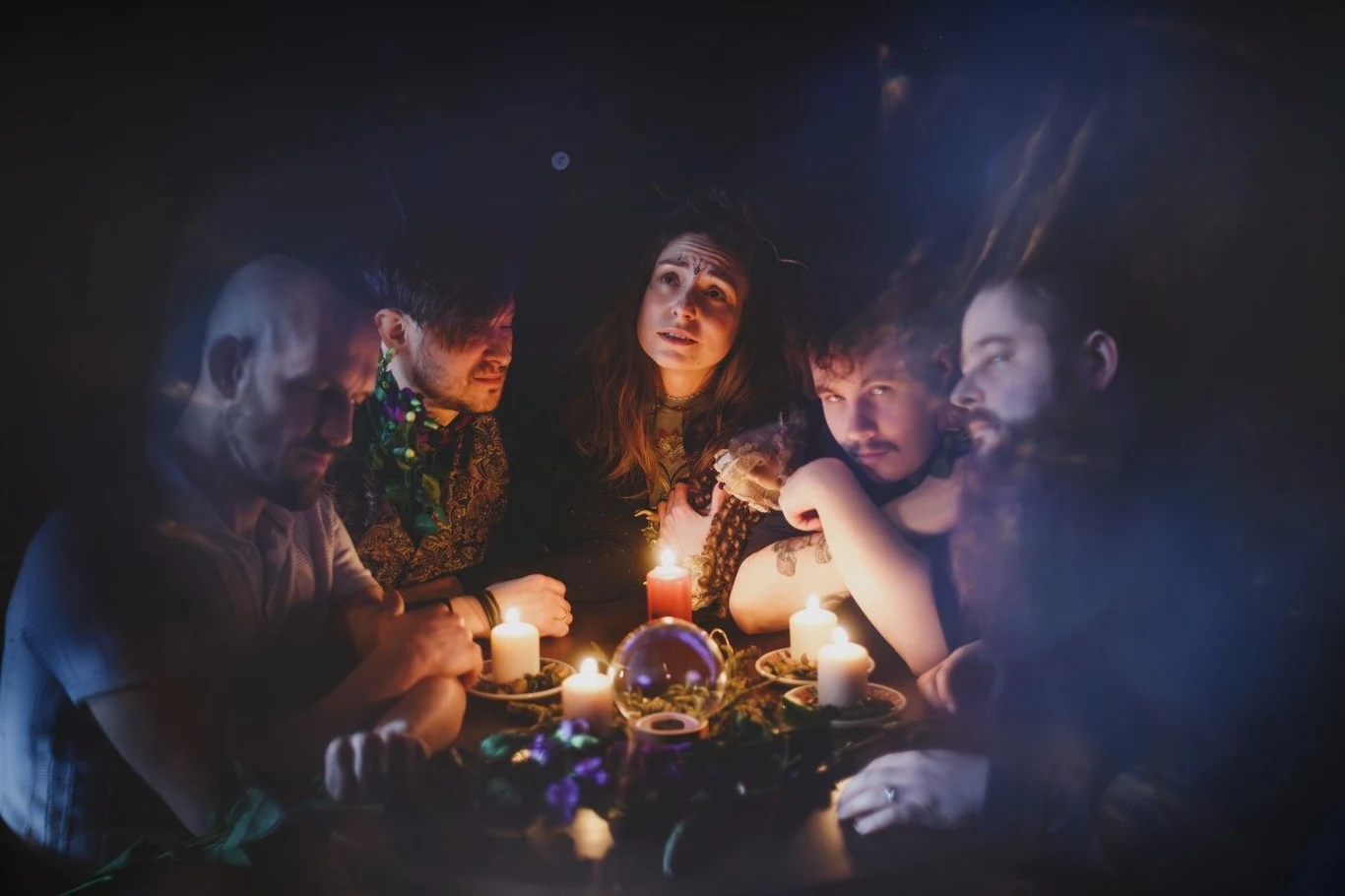 Źrenice szykują debiutancki album Grupa ludzi siedzi wokół stołu oświetlonego świecami, pośrodku znajduje się kryształowa kula oraz różne magiczne rekwizyty, atmosfera tajemnicza i pełna skupienia, każda osoba skoncentrowana na rytuale lub seansie spirytystycznym.