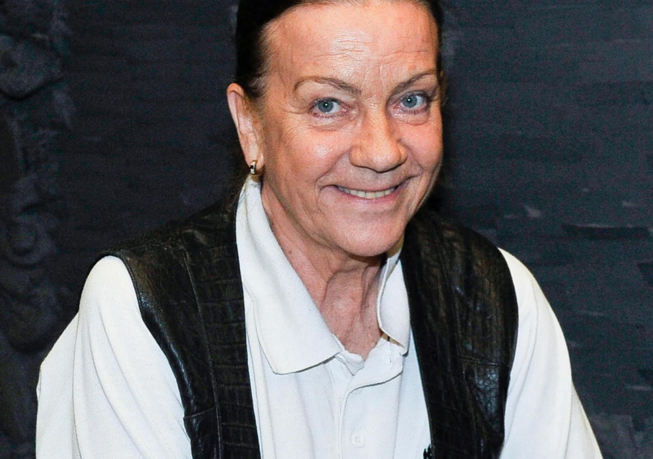 Barbara Burska Uśmiechnięta kobieta w białej koszuli polo oraz czarnym skórzanym bezrękawniku, z ciemnymi włosami spiętymi do tyłu, na ciemnym tle.