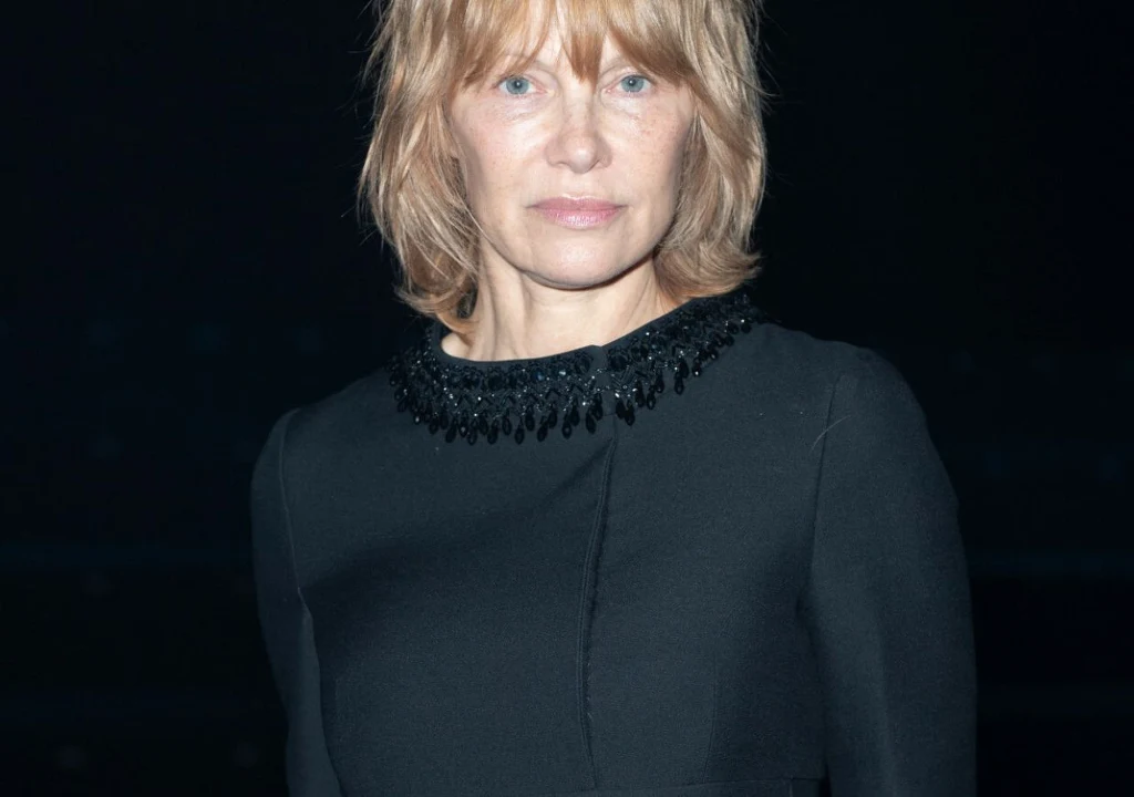 Pamela Anderson Kobieta o jasnych włosach sięgających ramion w czarnej sukni z ozdobnym kołnierzem, patrząca wprost na aparat na ciemnym tle.