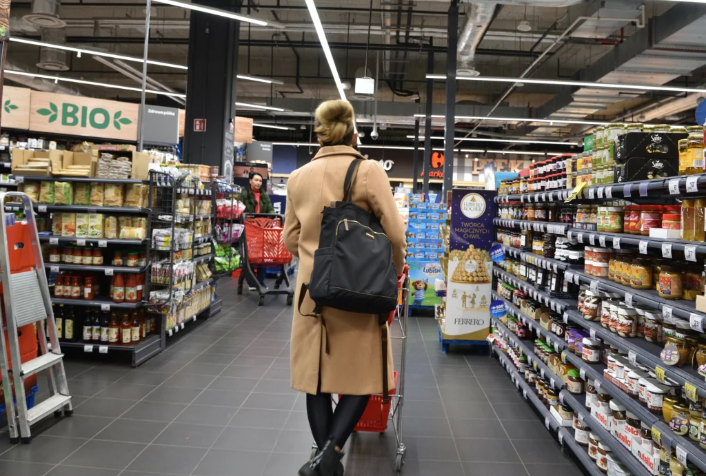 Carrefour w rękach państwowego koncernu? Kobieta w beżowym płaszczu i z czarnym plecakiem pcha wózek na zakupy alejką supermarketu, po bokach regały z produktami spożywczymi, w tle inni klienci wybierający towary.