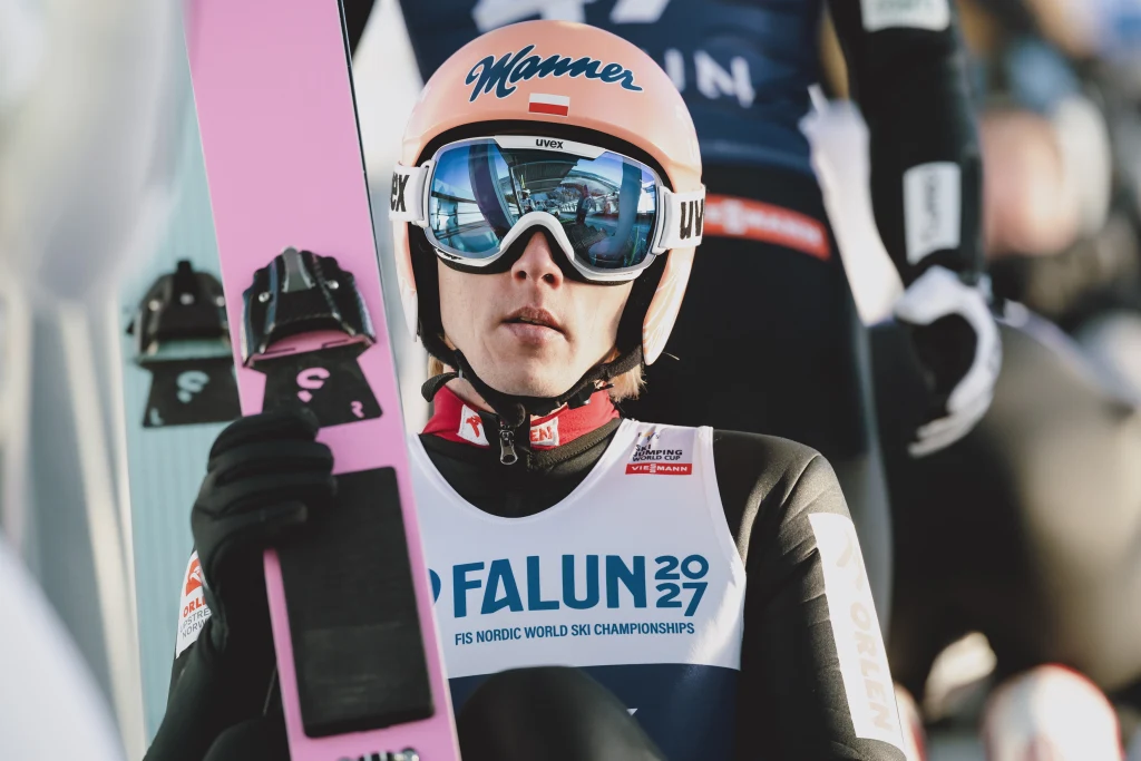 Dawid Kubacki Zawodnik skoków narciarskich w sportowym kombinezonie i kasku z goglami, trzymający narty, przygotowuje się do startu podczas mistrzostw świata w Falun 2027. Wyraz skupienia na twarzy, w tle inne osoby związane z wydarzeniem.