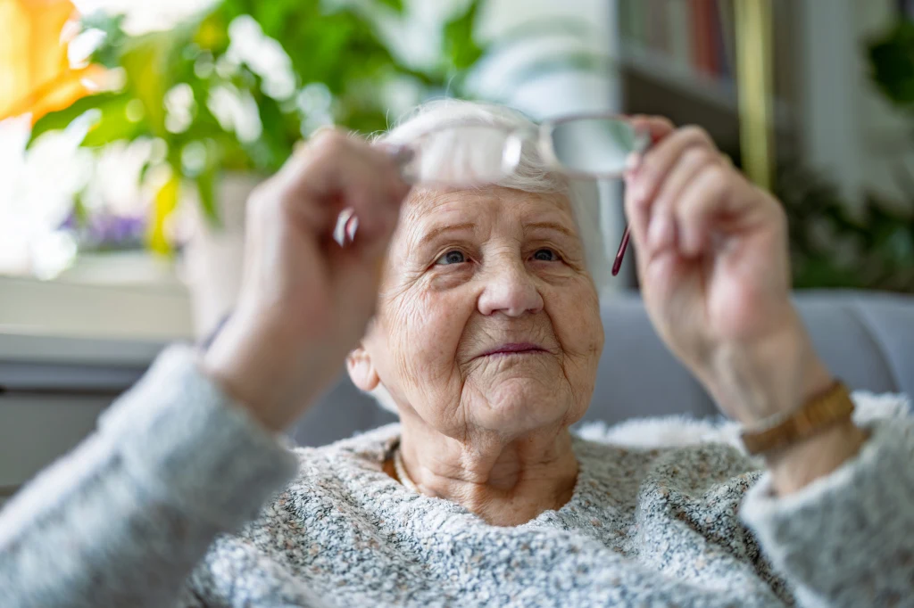 Złe nawyki, niezdrowa dieta i izolacja społeczna po cichu przyspieszają demencję Starsza kobieta o siwych włosach podnosi okulary, siedząc w jasnym pokoju z roślinami w tle.