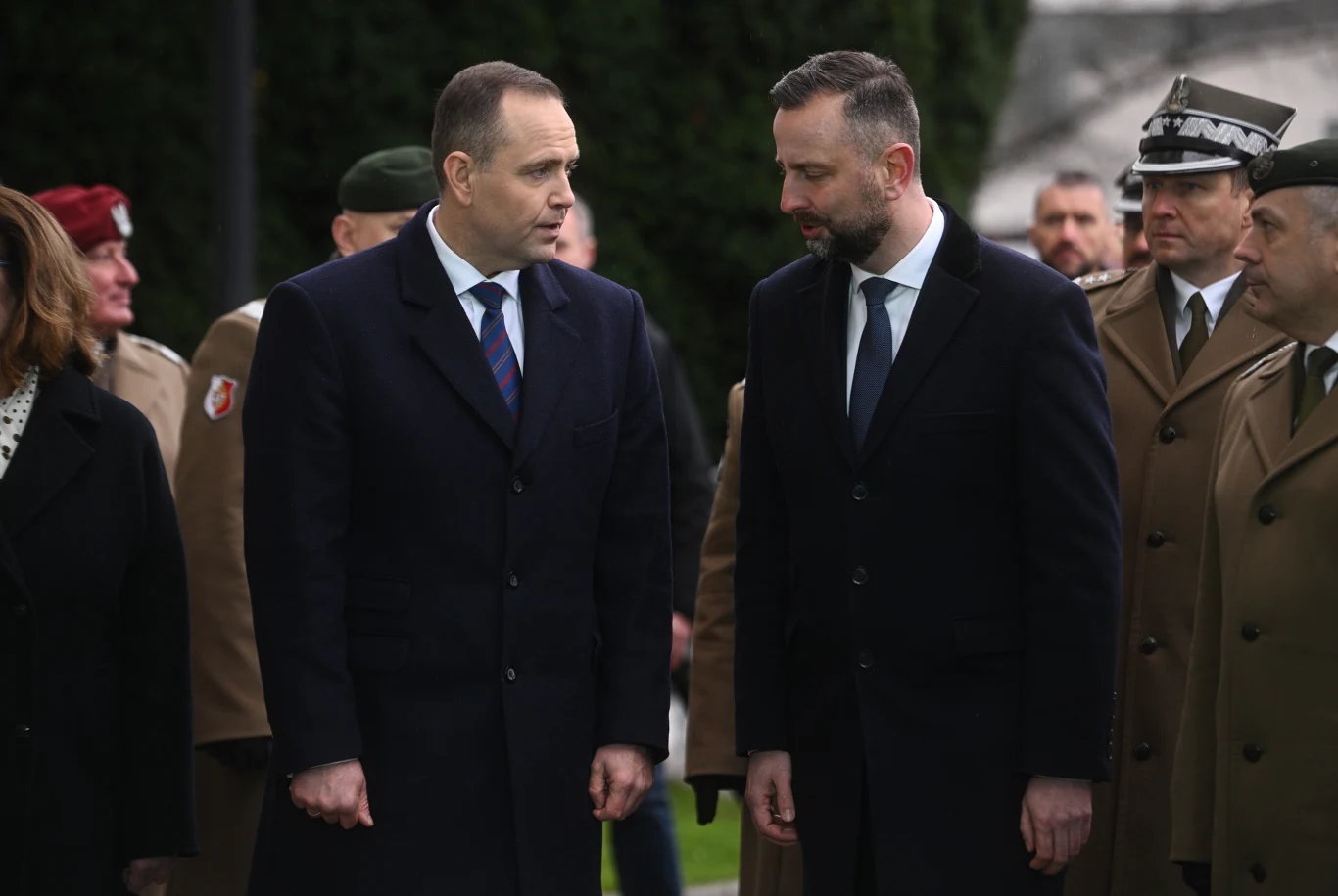 Spór wokół nominacji oficerskich trwa. Szef MON zapowiedział kolejne kroki Dwóch mężczyzn w garniturach stoi obok siebie na świeżym powietrzu w otoczeniu żołnierzy w mundurach oraz innych osób. W tle widoczne są drzewa i fragment budynku.