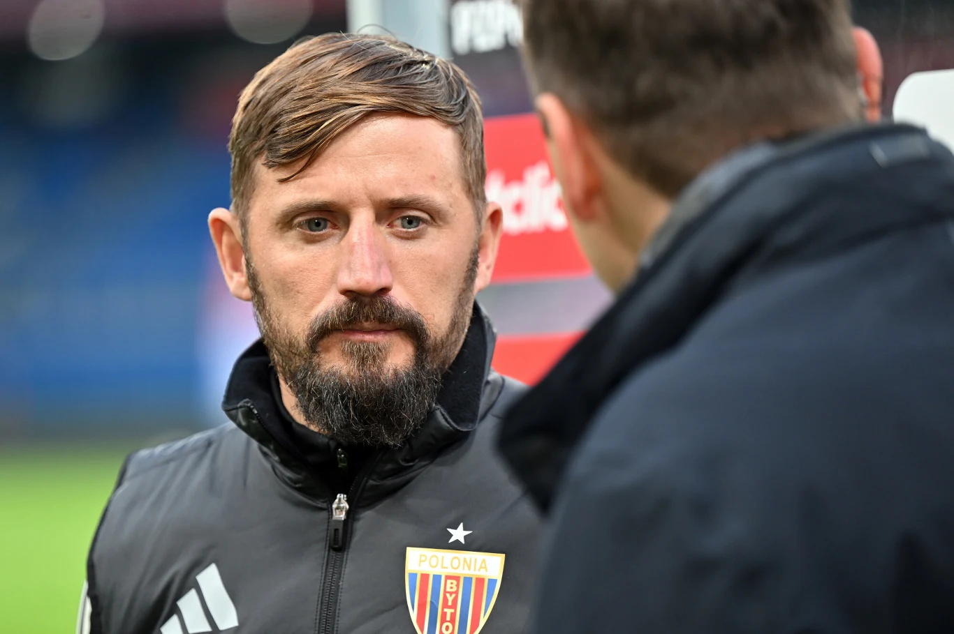 Łukasz Tomczyk Mężczyzna z krótką fryzurą i brodą w sportowej kurtce z emblematem klubu Polonia Bytom, stojący naprzeciw drugiej osoby, która widoczna jest od tyłu i rozmawiający z nią na tle rozmytego stadionu.