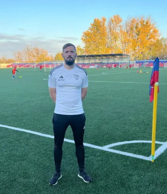 Łukasz Tomczyk Mężczyzna w białej sportowej koszulce i czarnych spodniach stoi przy chorągiewce narożnej na boisku piłkarskim, w tle widać innych graczy i trybuny oraz drzewa w jesiennych barwach.