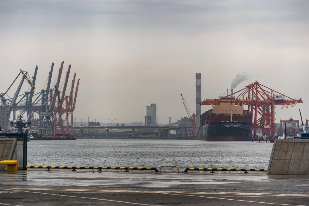 Port przemysłowy z licznymi dźwigami i kontenerowcem przy nabrzeżu, nad wodą unosi się lekka mgła, a nad jednym ze statków widać kłąb dymu.