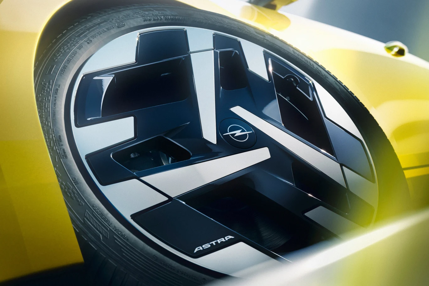 Felga samochodowa marki Opel z napisem Astra, na tle żółtego nadwozia pojazdu, zwracająca uwagę designerskim, geometrycznym wzorem.