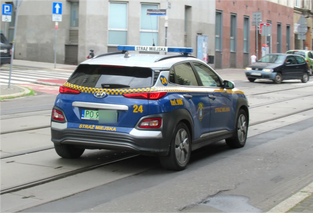 Pojazd Straży Miejskiej Hyundai Kona poruszający się po ulicy miasta, w tle nowoczesna zabudowa oraz piesi na chodniku.