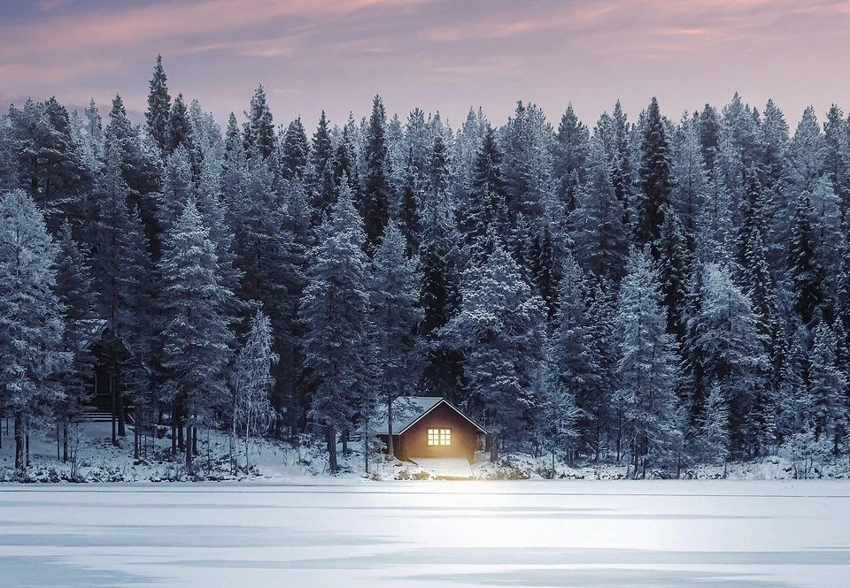 Mały drewniany domek z rozświetlonymi oknami stoi na skraju zaśnieżonego lasu, otoczony wysokimi sosnami. Przed domkiem zamarznięte jezioro, a niebo przybiera fioletowo-różowe odcienie zmierzchu.