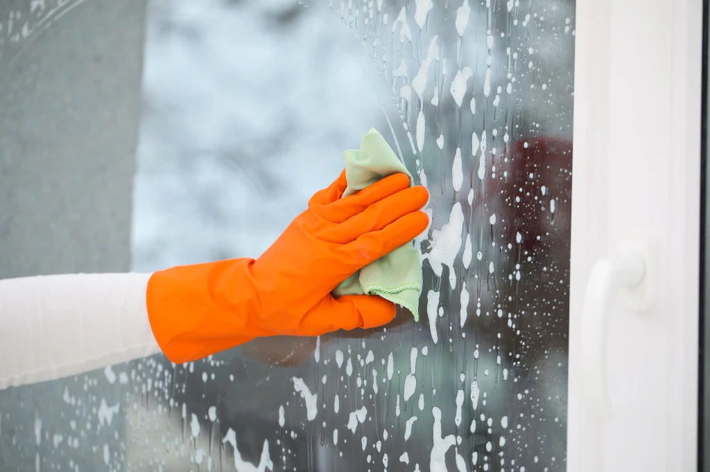 Czy mycie okien przed świętami to dobry pomysł? Ręka w pomarańczowej gumowej rękawicy ściera szyby okienne ściereczką, na powierzchni okna widoczna jest piana po środku czyszczenia.