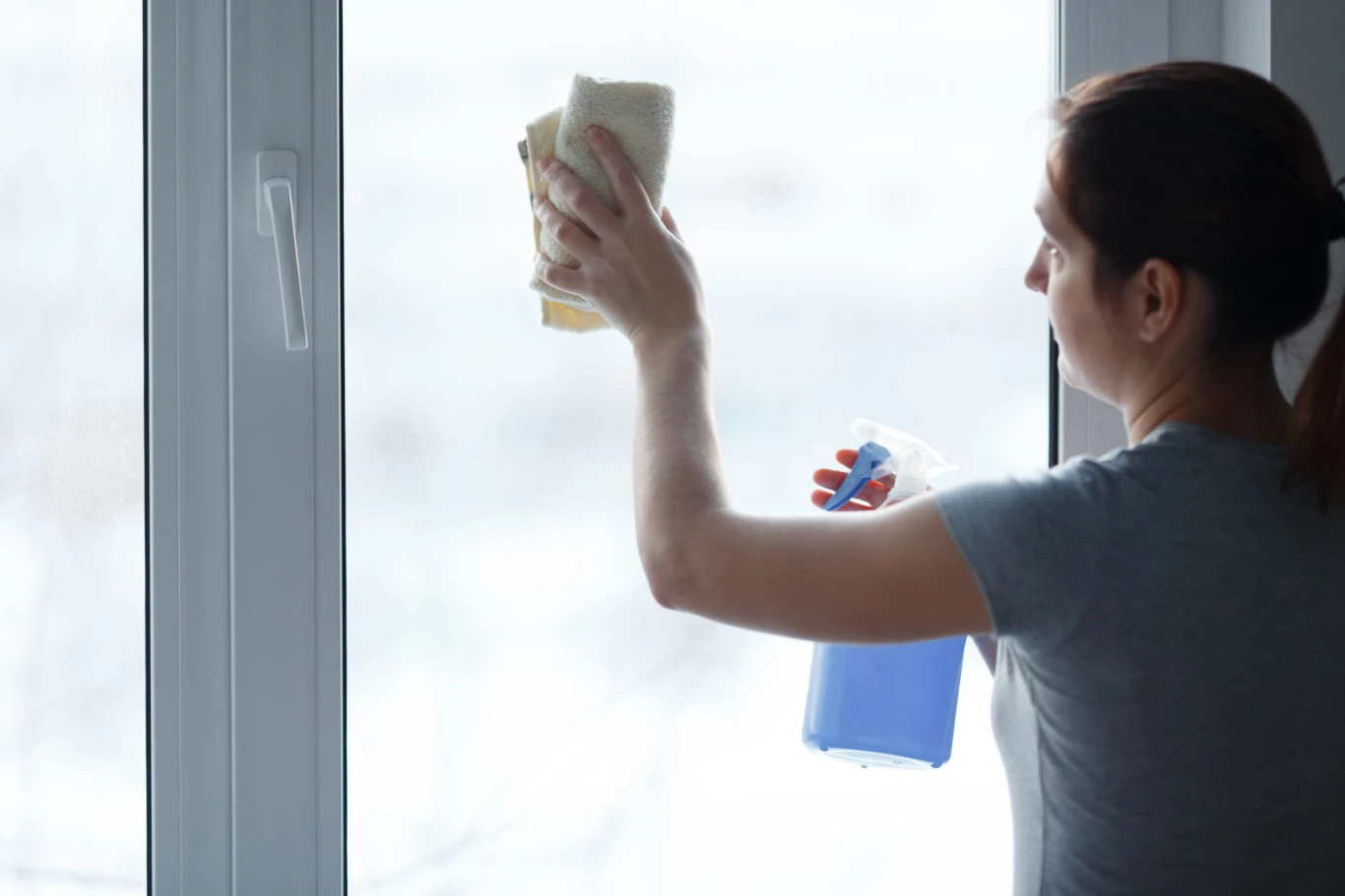 Kobieta czyści szybę okienną za pomocą niebieskiego płynu i ściereczki, w tle jasne, niewyraźne otoczenie na zewnątrz.