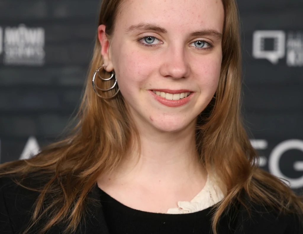 Młoda kobieta z długimi, jasnymi włosami i niebieskimi oczami, ubrana w czarną marynarkę oraz kremową bluzkę, delikatnie uśmiechnięta. W tle rozmazana ściana z ciemnymi napisami.