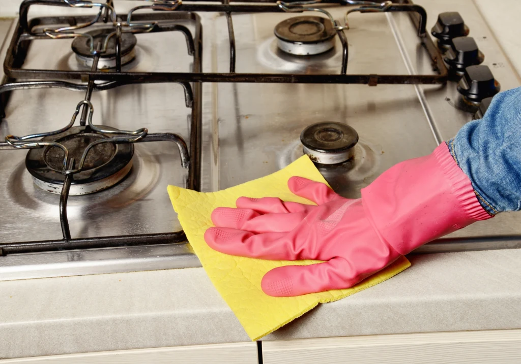 Ręka w różowej gumowej rękawiczce czyści kuchenkę gazową żółtą gąbką. Kuchenka posiada metalowe ruszty i pokrętła, powierzchnia jest starannie przecierana.
