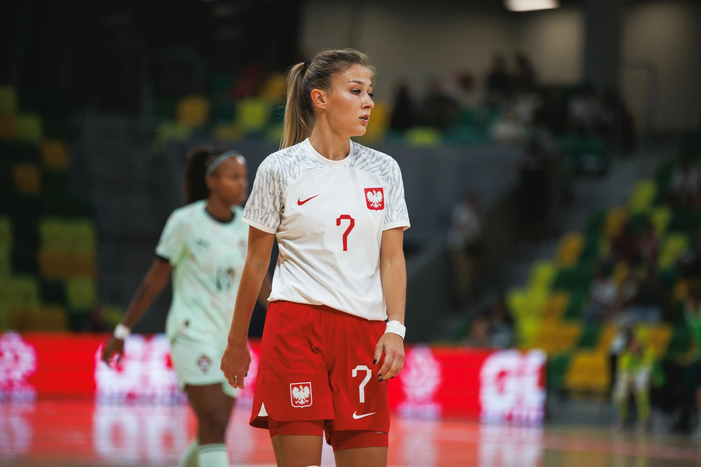 Agata Bała Piłkarka w białej koszulce i czerwonych spodenkach z orłem na piersi oraz numerem 7 stoi na hali sportowej, skupiona na grze, w tle rozmyte trybuny oraz inna zawodniczka.