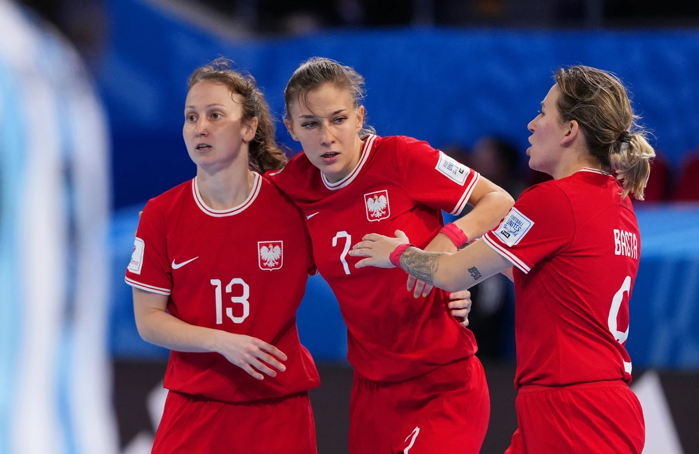 Reprezentacja Polski w futsalu Trzy kobiety w czerwonych koszulkach reprezentacji Polski podczas meczu piłki ręcznej. Jedna z zawodniczek wspiera drugą, która wygląda na skupioną lub zaniepokojoną. W tle widoczny fragment boiska oraz sylwetka zawodniczki przeciwnej drużyny.