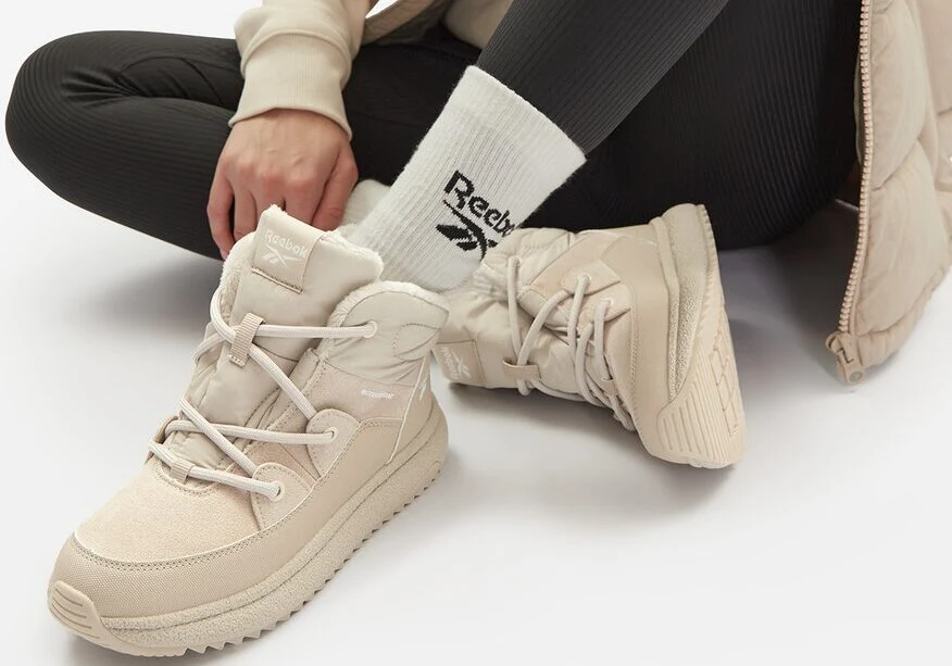 Wygodne śniegowce na zimę Beżowe buty zimowe na grubej podeszwie, noszone przez osobę ubraną w czarne legginsy, jasne skarpety Reebok oraz jasną kurtkę. Osoba siedzi z nogą założoną na nogę, ukazując szczegóły obuwia i sportowego stylu.