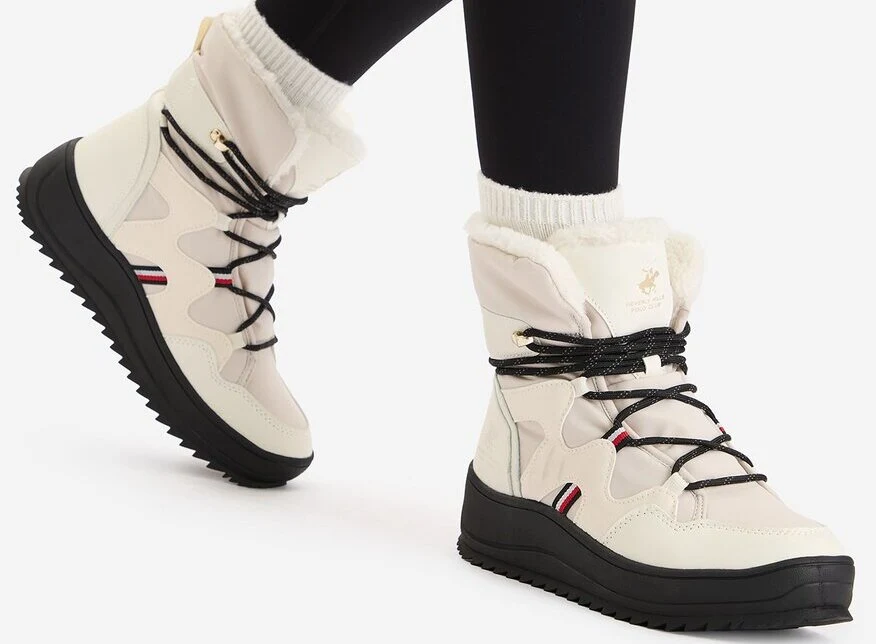 Białe zimowe buty z czarną podeszwą i czarnymi sznurówkami, ozdobione małymi czerwono-biało-granatowymi detalami. Osoba nosi ciemne legginsy, a buty mają miękkie, futrzane wykończenie przy kostce.