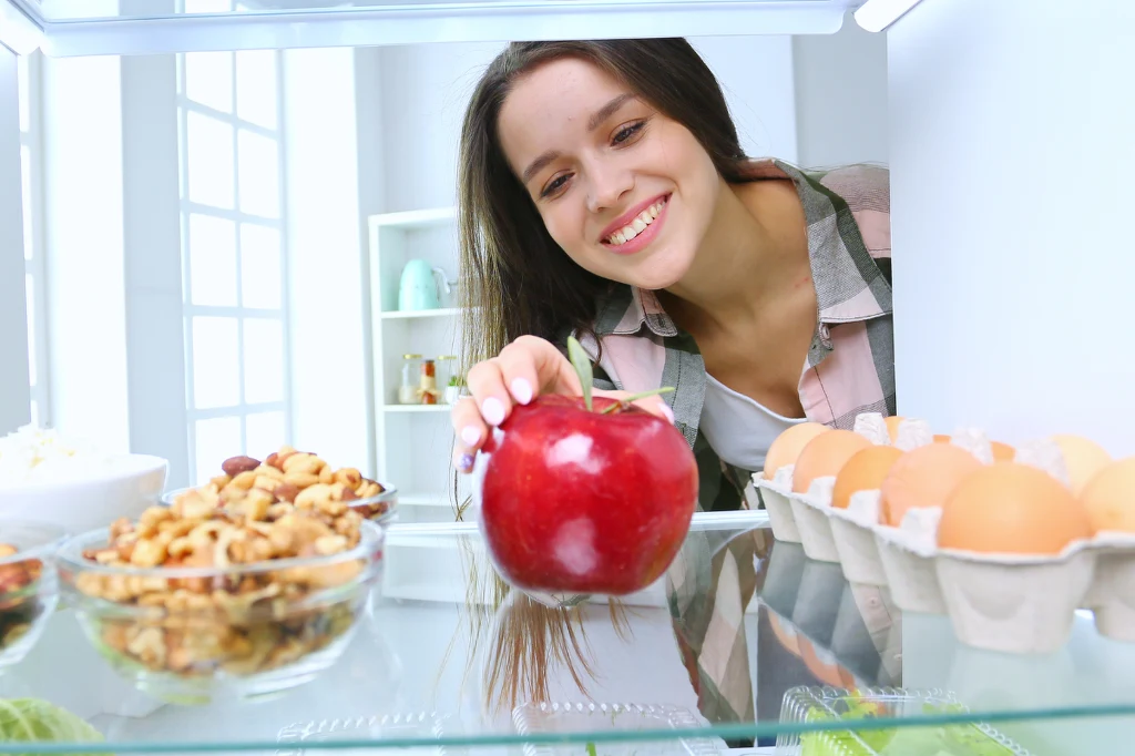 Młoda kobieta sięga po duże czerwone jabłko znajdujące się w lodówce, obok widoczne są jajka oraz miseczki z orzechami i innymi przekąskami.