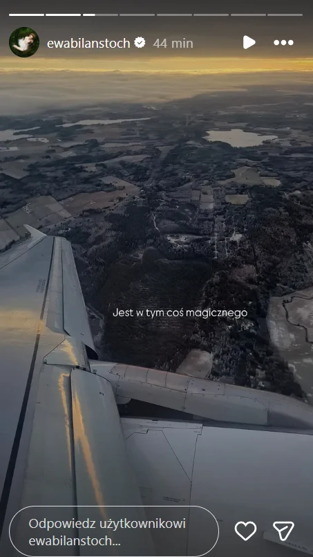 Widok skrzydła samolotu podczas lotu nad rozległym krajobrazem z polami, lasami i jeziorami, w tle widoczne zachmurzone niebo i delikatne światło zachodzącego słońca.