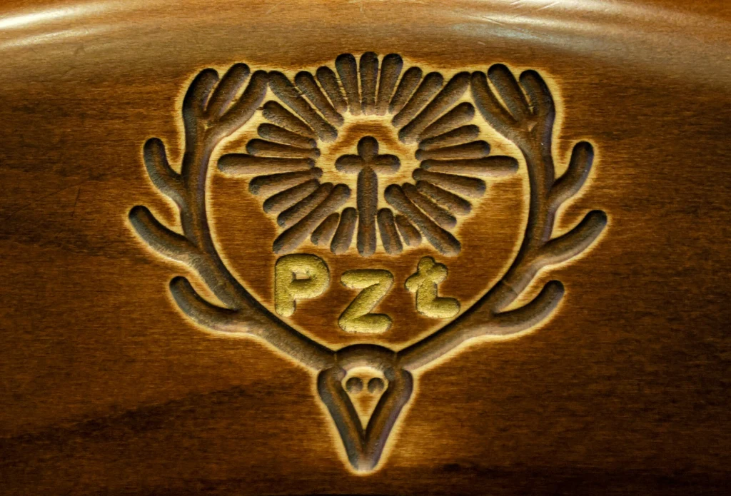 Grawerunek w drewnie przedstawiający stylizowane poroże, w jego wnętrzu znajduje się krzyż, rozchodzące się promienie oraz litery PZŁ podkreślone zlotą farbą.