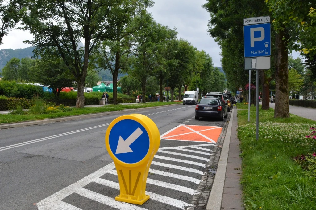 Kto może parkować na pomarańczowych kopertach bez narażania się na mandat? /Urząd Miasta Zakopane Płatny parking przy ulicy z wyznaczonymi miejscami postojowymi, znakiem poziomym zakazującym postoju na części strefy oraz dużym żółtym znakiem wskazującym kierunek ruchu. W tle drzewa i kilka zaparkowanych samochodów.