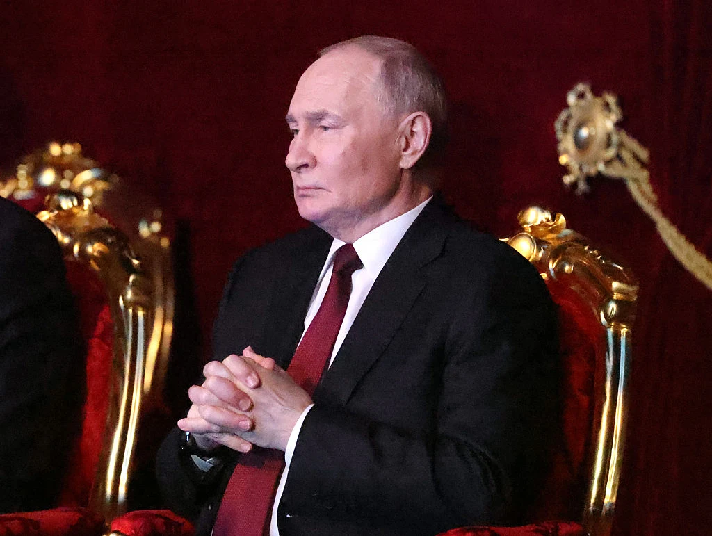 Mężczyzna w garniturze siedzi na bogato zdobionym, złotym fotelu na tle czerwonych zasłon, dłonie splecione i spokojny wyraz twarzy.