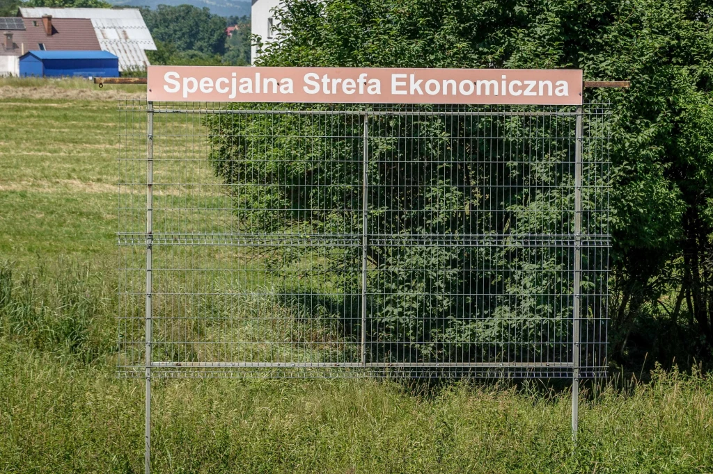 Metalowa konstrukcja ogrodzeniowa z napisem 'Specjalna Strefa Ekonomiczna' ustawiona na tle zielonego pola i drzew. W oddali widoczne fragmenty budynków wiejskich.