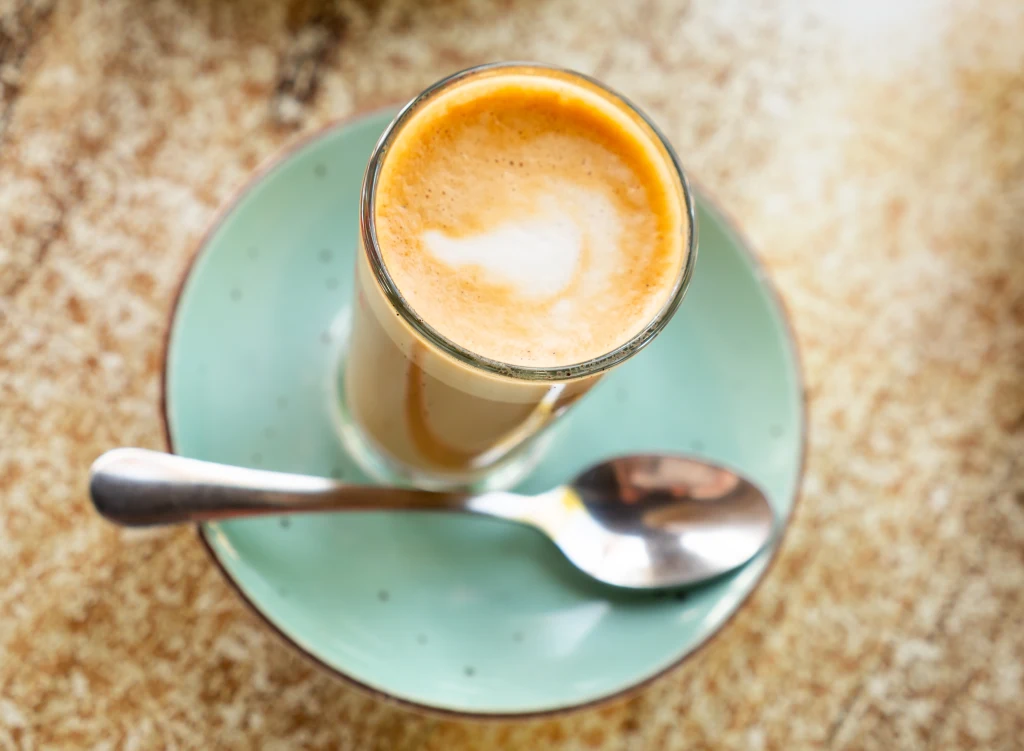 Cortado to idealny balans między mocnym espresso a słodkim cappuccino Szklanka kawy z mleczną pianką ustawiona na błękitnym spodku, obok leży metalowa łyżeczka. Tło stanowi jasny, marmurkowy blat stołu.