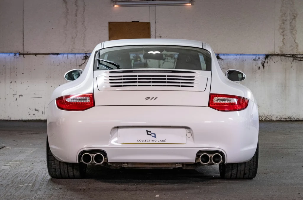 Tył białego samochodu sportowego Porsche 911 z widocznymi czterema końcówkami wydechu, czerwonymi światłami tylnimi oraz charakterystycznym logo 911. Auto stoi w garażu z białymi, częściowo zabrudzonymi ścianami.