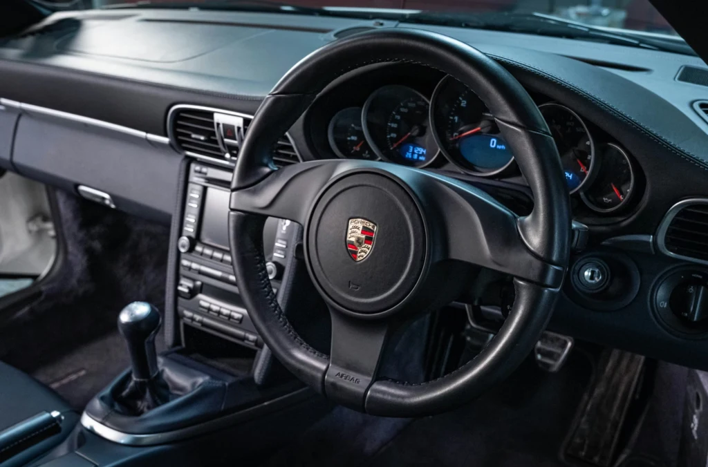 Wnętrze samochodu sportowego marki Porsche z kierownicą po prawej stronie, widocznym zestawem wskaźników, manualną skrzynią biegów oraz centralnym panelem sterowania.