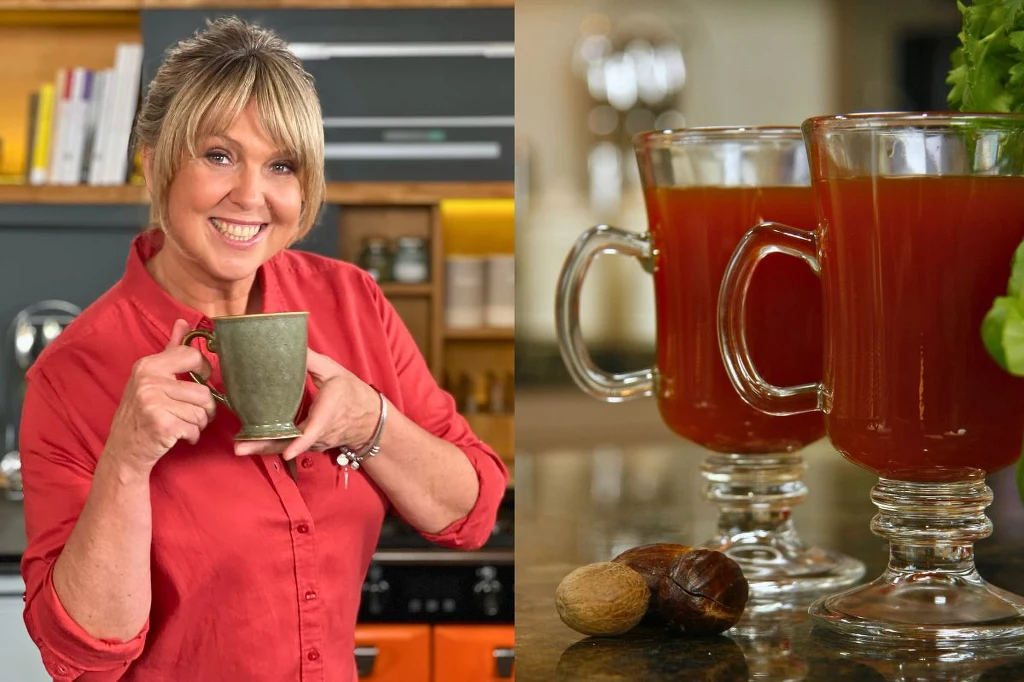 Herbata zimowa Uśmiechnięta kobieta ubrana w czerwoną koszulę trzyma kubek, obok na stole znajdują się dwie szklanki z czerwonym napojem oraz całe orzechy i świeże zioła na blacie kuchennym.