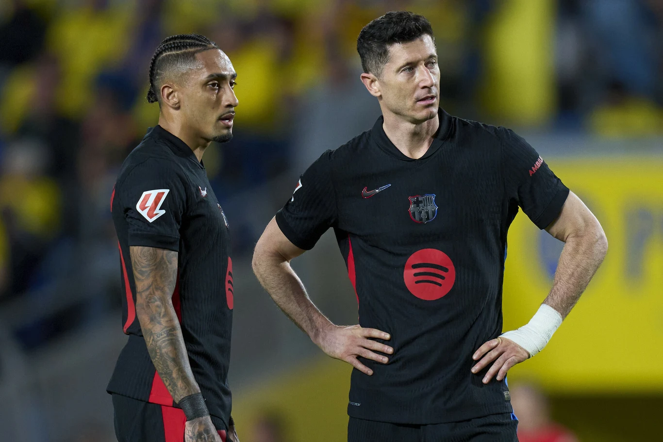 Dwóch piłkarzy w koszulkach FC Barcelony stoi na boisku podczas meczu, jeden z nich ma ręce na biodrach, obaj są skupieni i patrzą w jednym kierunku, w tle widać rozmyte sylwetki oraz żółte barwy trybun.