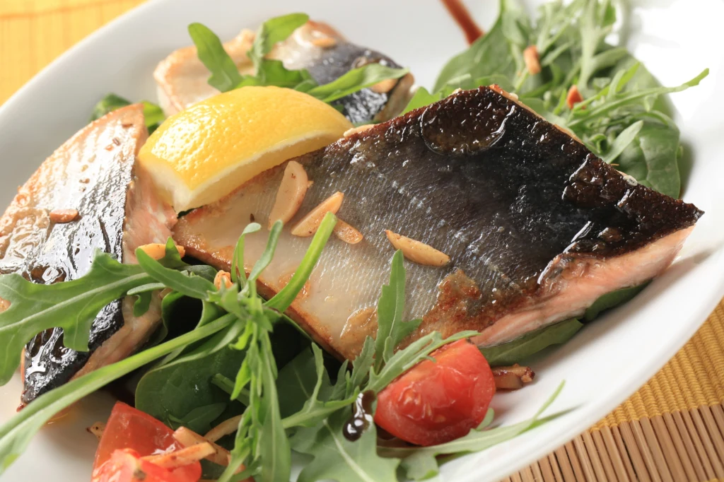 Pstrąg tęczowy jest bogaty w wielonienasycone kwasy tłuszczowe Kawałki grillowanej ryby na rukoli z cząstką cytryny, prażonymi migdałami i pomidorkami koktajlowymi, polane lekko sosem.