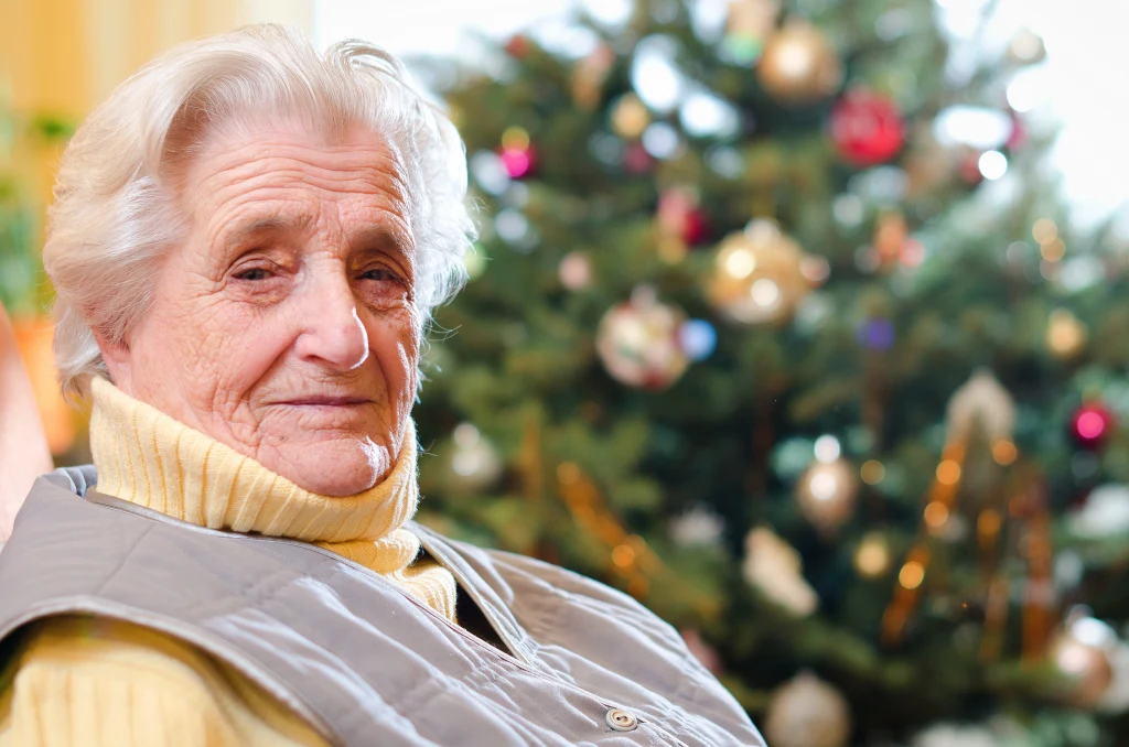 Sprawdź swój horoskop na grudzień od wróżki Mariwy Starsza kobieta w jasnym swetrze siedzi przed udekorowaną choinką ze złotymi i czerwonymi bombkami, w tle widoczne świąteczne światełka.